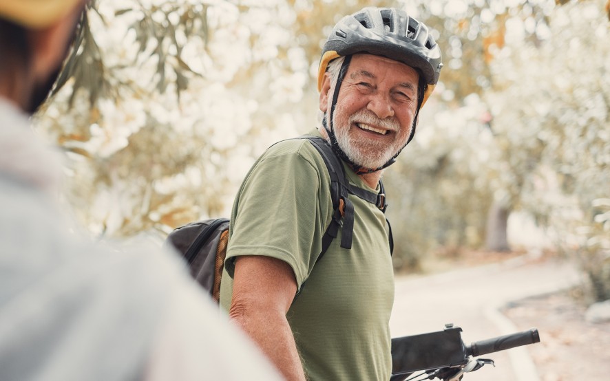 Uomo anziano in bicicletta con il casco e la zaino. Nell’articolo parliamo delle raccomandazioni per fare ciclismo in sicurezza dopo i 70 anni